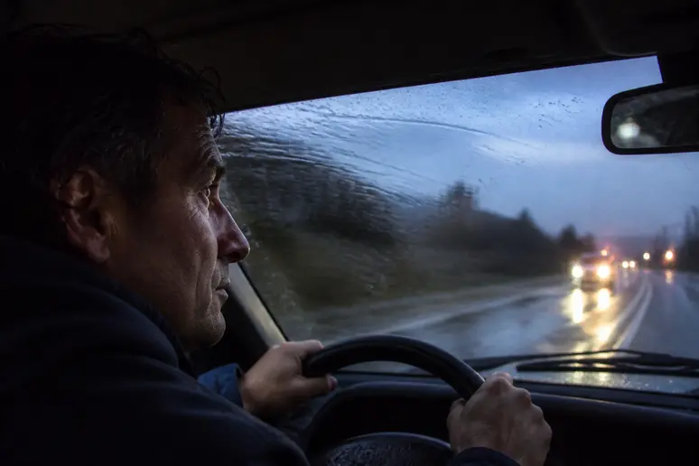 Homme conduisant sous la pluie, visibilité réduite par les essuie-glaces usés, inquiétude sur son visage.